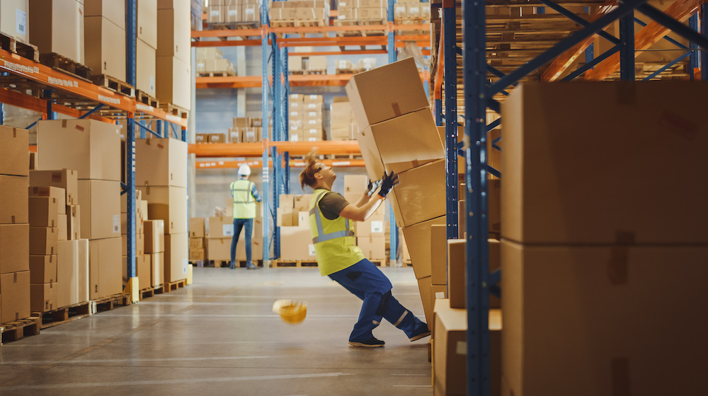 Shot of a Warehouse Worker Has Work Related Accident. He is Falling Down BeforeTrying to Pick Up Heavy Cardboard Box from the Shelf. Hard Injury at Work.